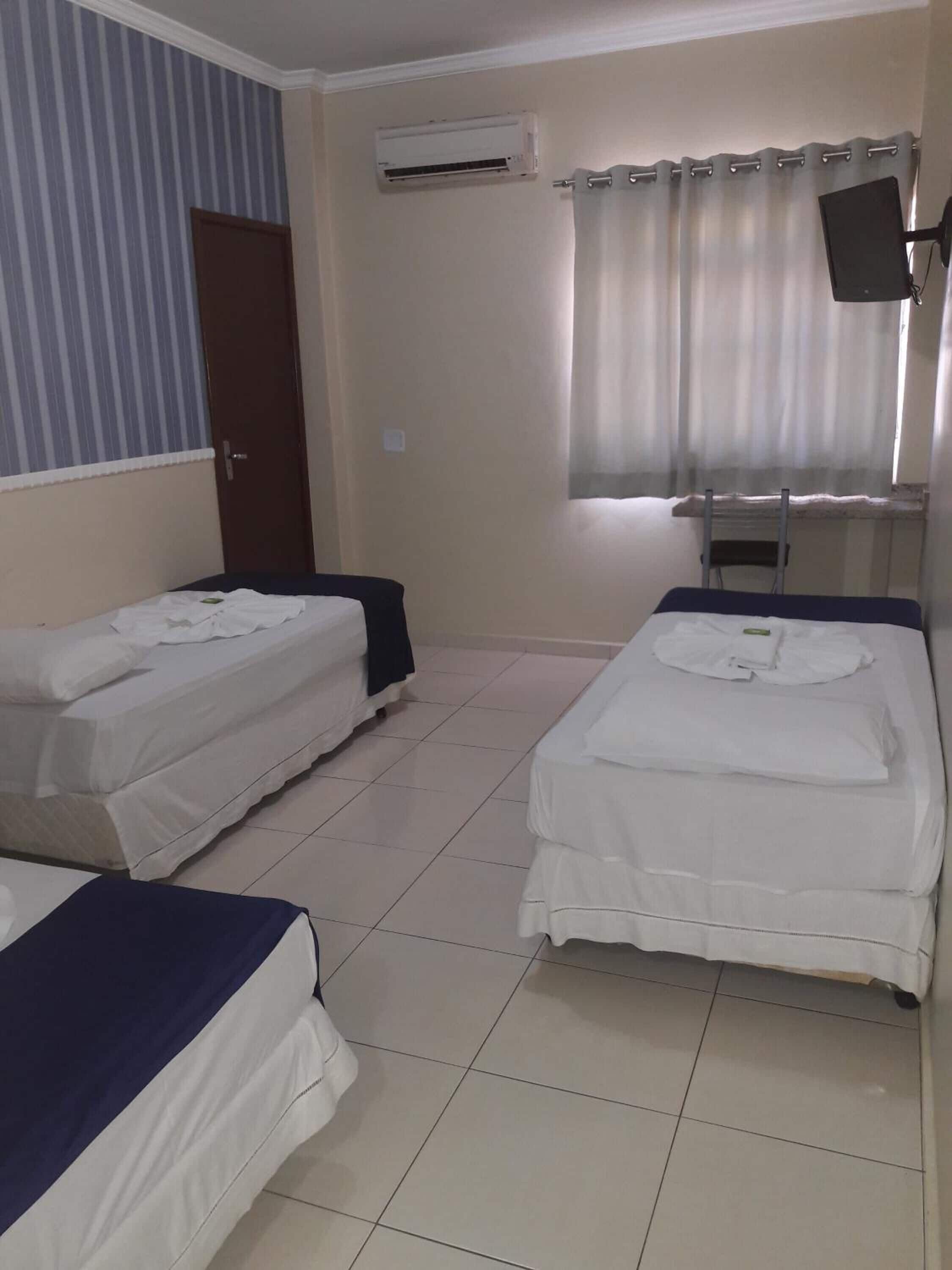 Quarto com duas camas, uma porta, ar condicionado, janela com cortinas e televisão na parede.