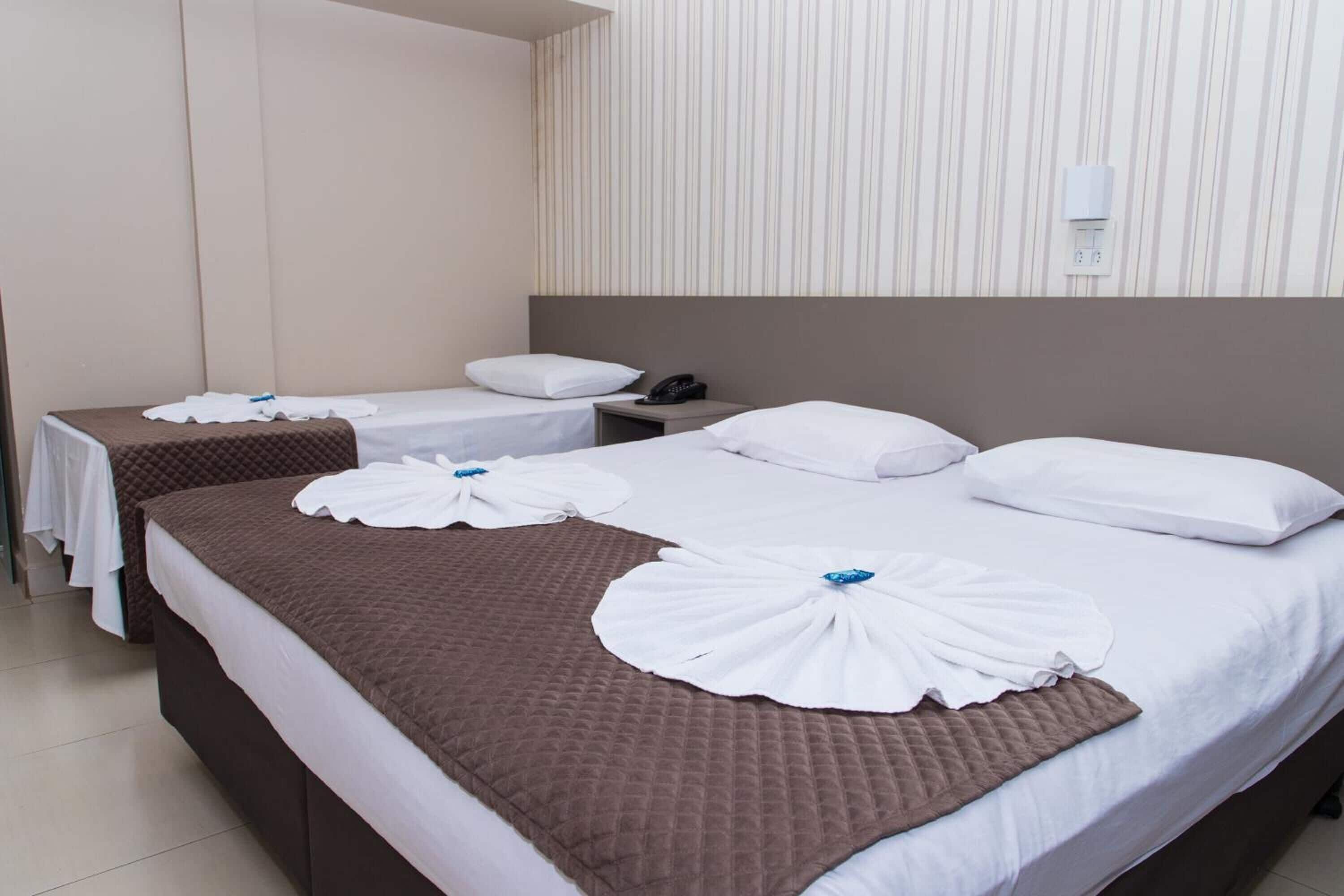 A hotel room with two beds, each with a brown quilted bedspread and white linens. There are white towels arranged on the beds, and a white wall-mounted phone.