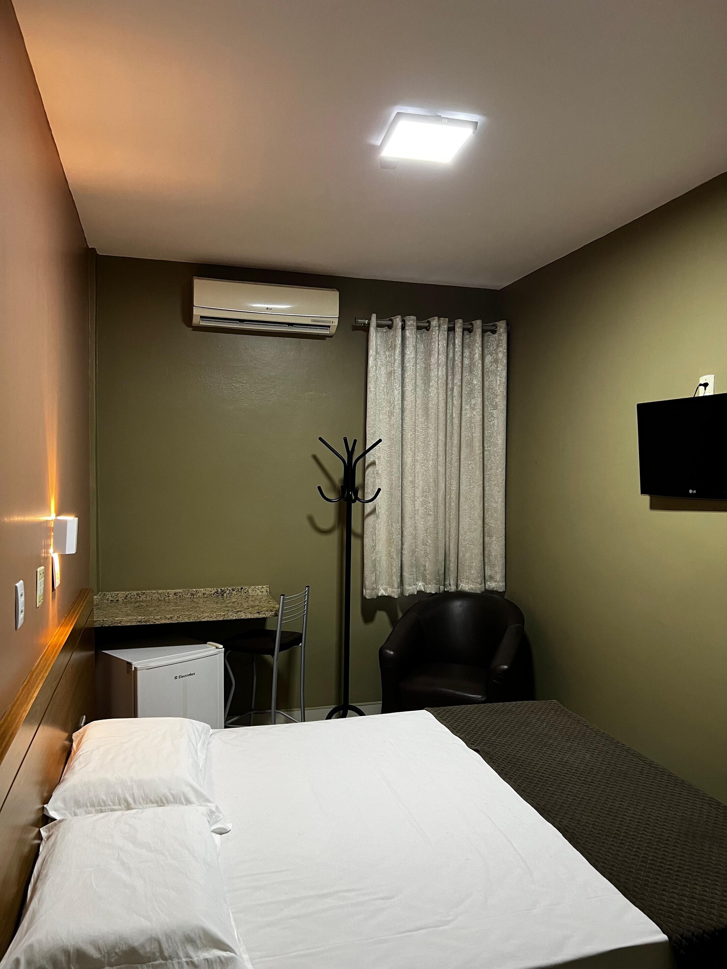 A hotel room with a bed, a desk, a chair, an air conditioner, and a television.