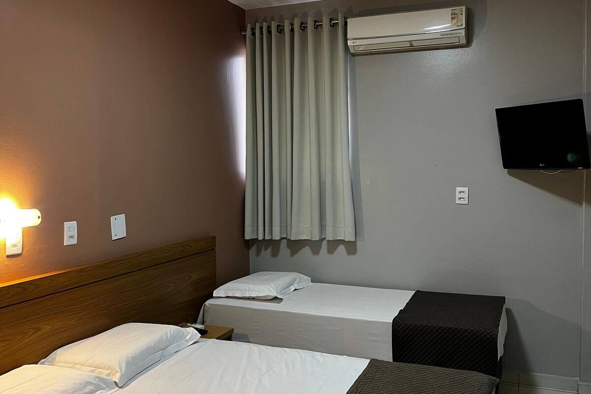 Une chambre d’hôtel avec deux lits, un luminaire au plafond, un appareil de climatisation, une télévision et un rideau.