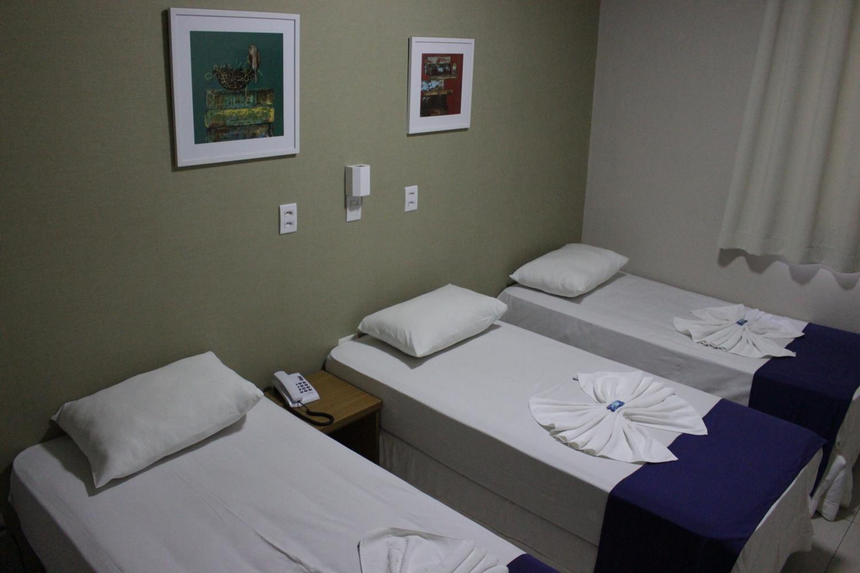 Habitación de hotel con tres camas individuales, cada una con una almohada blanca y una toalla blanca doblada. Hay una mesita de noche con un teléfono y un interruptor de luz en la pared. Dos cuadros enmarcados están colgados en la pared sobre las camas.