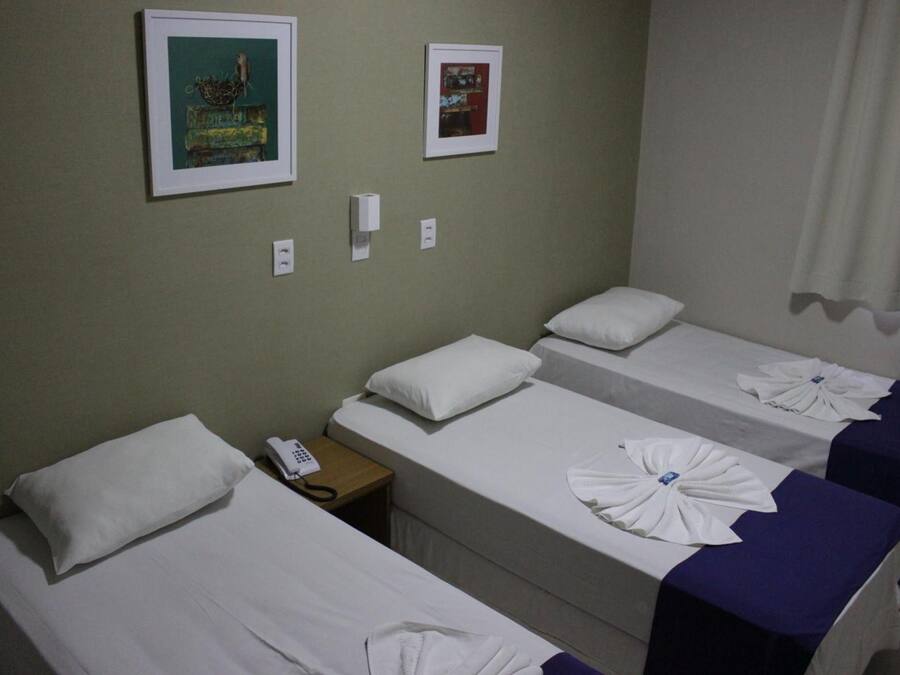 Habitación de hotel con tres camas individuales, cada una con una almohada blanca y una toalla blanca doblada. Hay una mesita de noche con un teléfono y un interruptor de luz en la pared. Dos cuadros enmarcados están colgados en la pared sobre las camas.