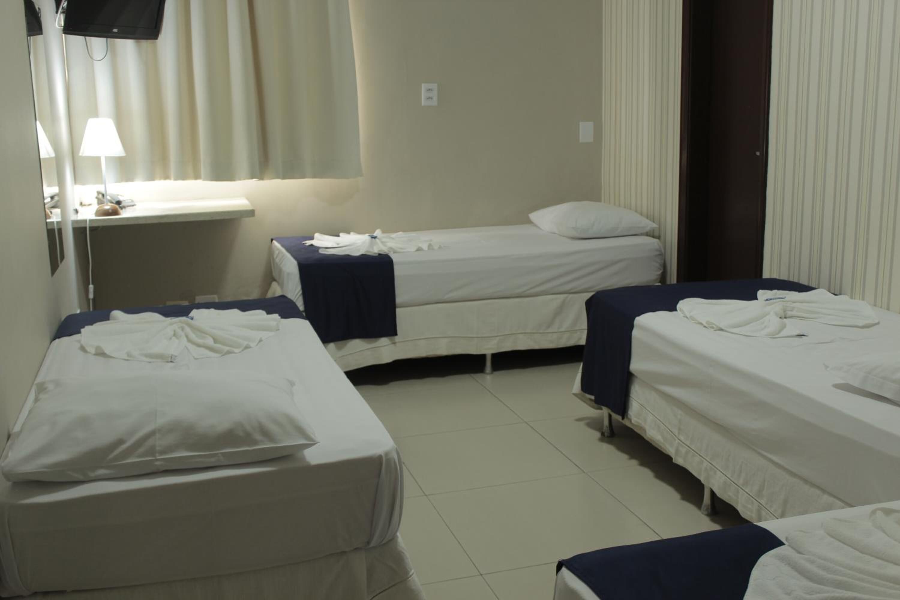 Quarto de hotel com três camas de solteiro, uma escrivaninha com luminária e uma televisão na parede.