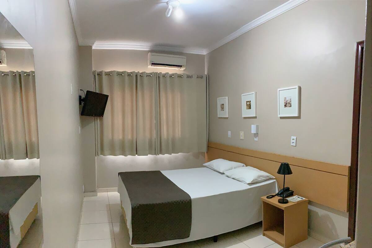 Une chambre d’hôtel avec un lit, une table de chevet, une lampe, une plante en pot et des tableaux encadrés au mur.