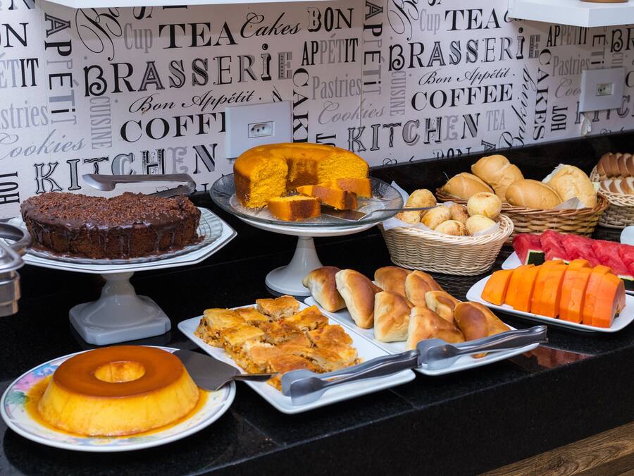 Buffet com diversos doces e pastelaria, incluindo um bolo de chocolate, um pudim e vários tipos de pão e pães de forma.