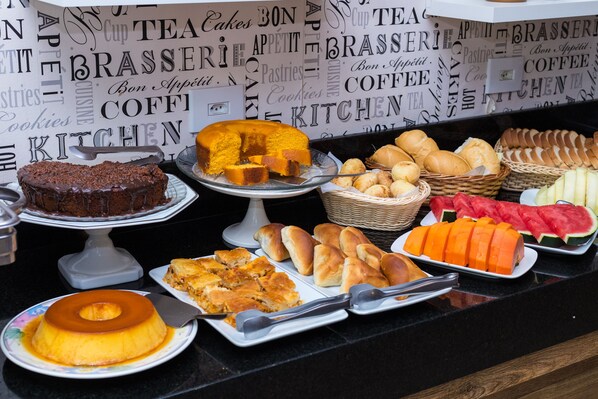 Un bufé con diversos postres y pasteles, que incluye un pastel de chocolate, un flan y varios tipos de pan y bollos.