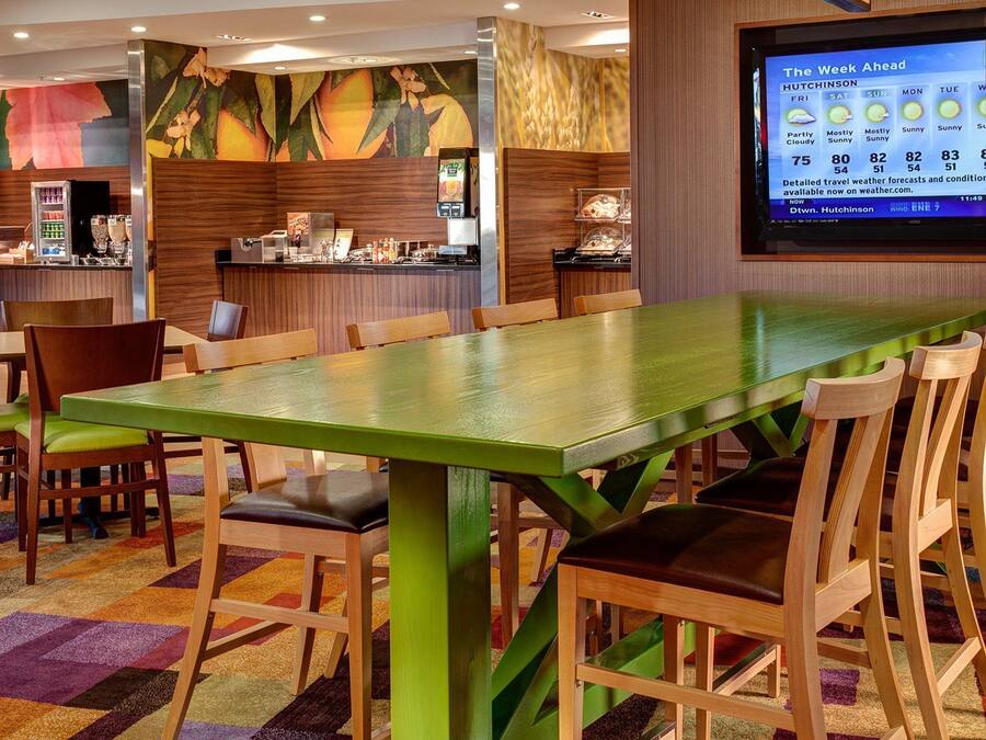 A hotel dining area with a long green table, wooden chairs, and a colorful carpet. A screen displaying weather information is visible in the background.