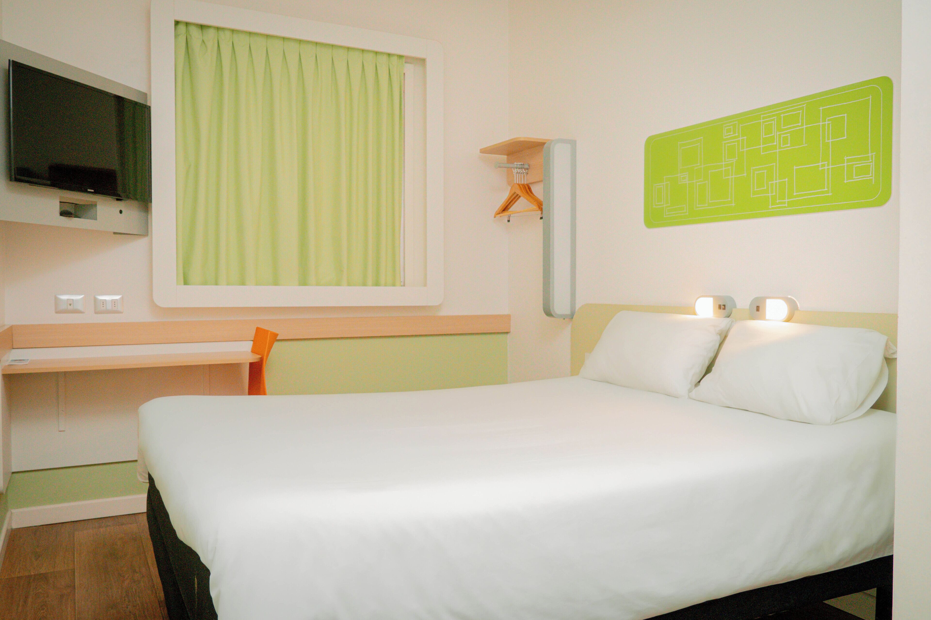 A hotel room with a bed, a desk, a television, and a wall-mounted light fixture.