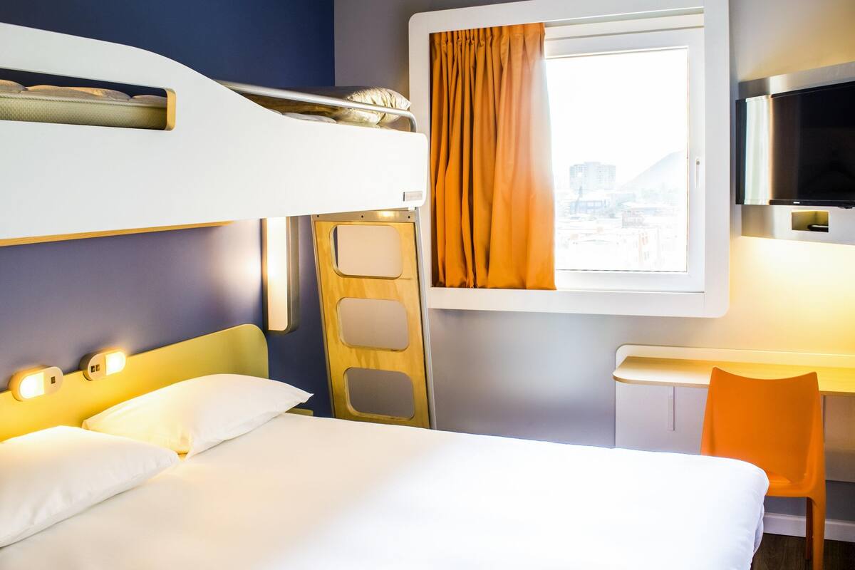 Une chambre d’hôtel avec un lit superposé, un bureau, une chaise et une fenêtre avec des rideaux.