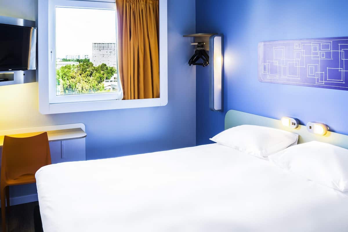 Une chambre d’hôtel équipée d’un lit, d’un bureau, d’une télévision et d’une fenêtre donnant sur un paysage verdoyant et des bâtiments.