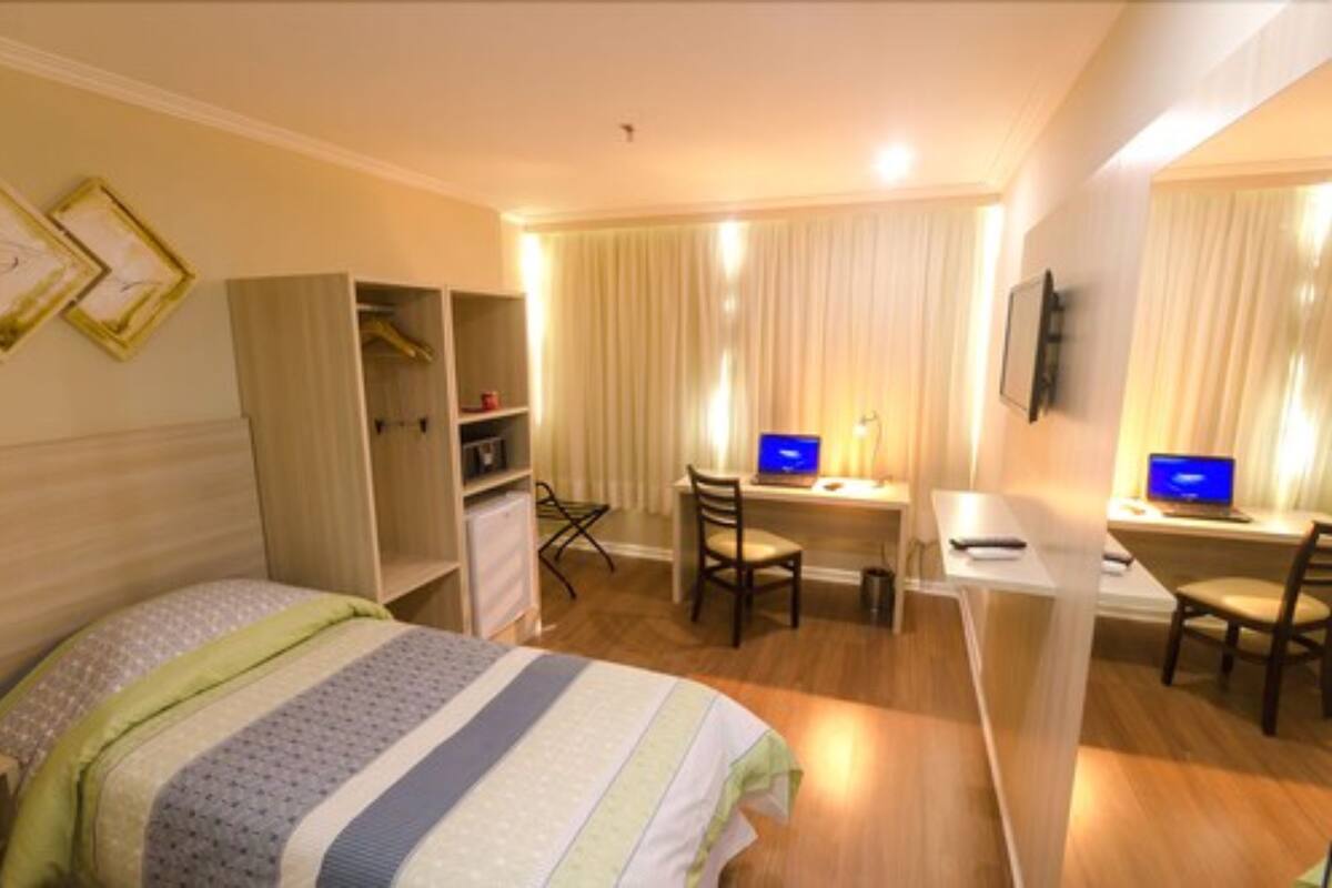 Ein Hotelzimmer mit einem Bett, einem Schreibtisch mit Computer, einem Stuhl, einem Kleiderschrank und einem an der Wand befestigten Fernseher.