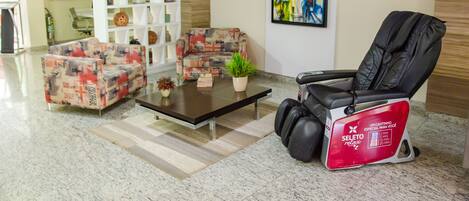 A lobby with two patterned sofas, a massage chair, a coffee table with a plant, and a bookshelf.