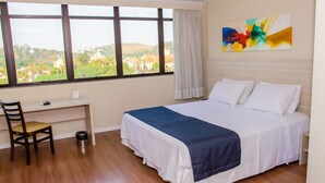 A hotel room with a large bed, a desk, and a chair. There is a window with a view of a cityscape. - Seleto Hotel (Volta Redonda)