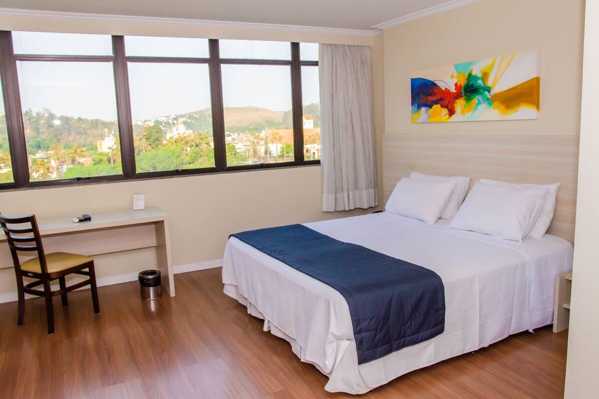 Ein Hotelzimmer mit einem großen Bett, einem Schreibtisch und einem Stuhl. Ein Fenster bietet Blick auf eine Stadtlandschaft.