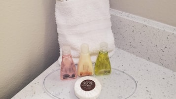A bathroom with a white towel hanging on a wall, a glass soap dish with three bottles of liquid soap, and a round soap dish with a bar of soap.