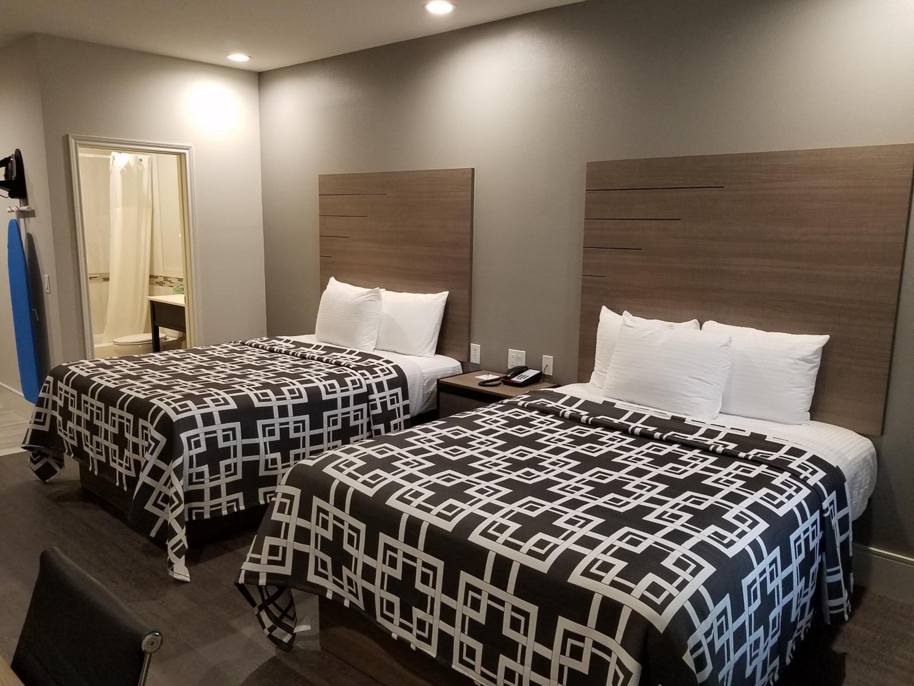 A hotel room with two beds, each with a patterned bedspread and white pillows. There is a bedside table with a phone and a lamp. The room has a wooden headboard and a door leading to a bathroom.