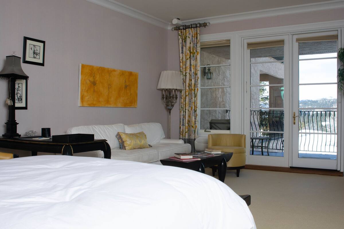 A hotel room with a bed, a sofa, a small table, a chair, a balcony with a railing, and a door with glass panels.