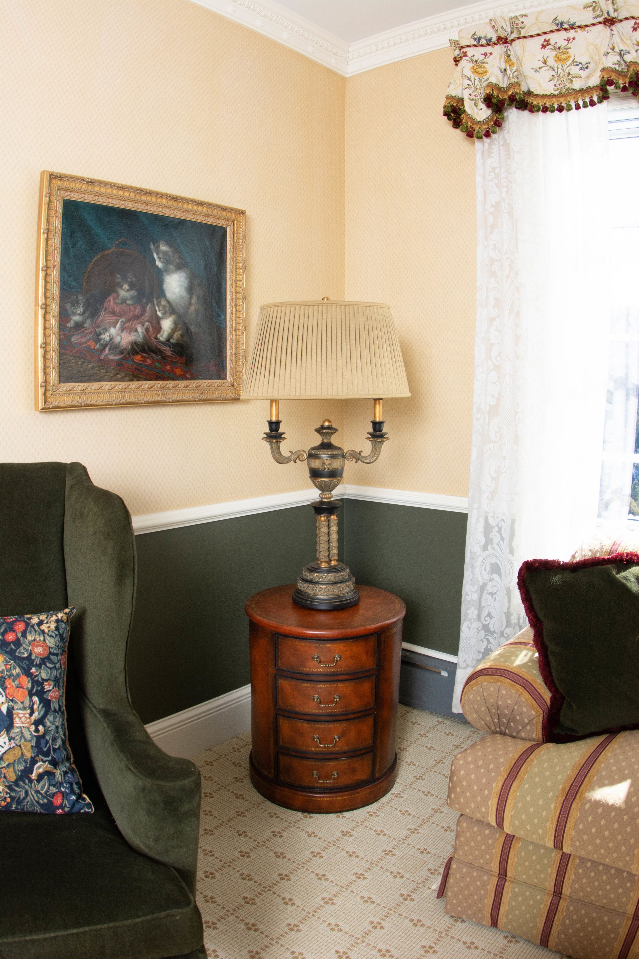 A room with a green armchair, a patterned sofa, a wooden side table with a lamp, a framed painting, and a window with a white curtain.