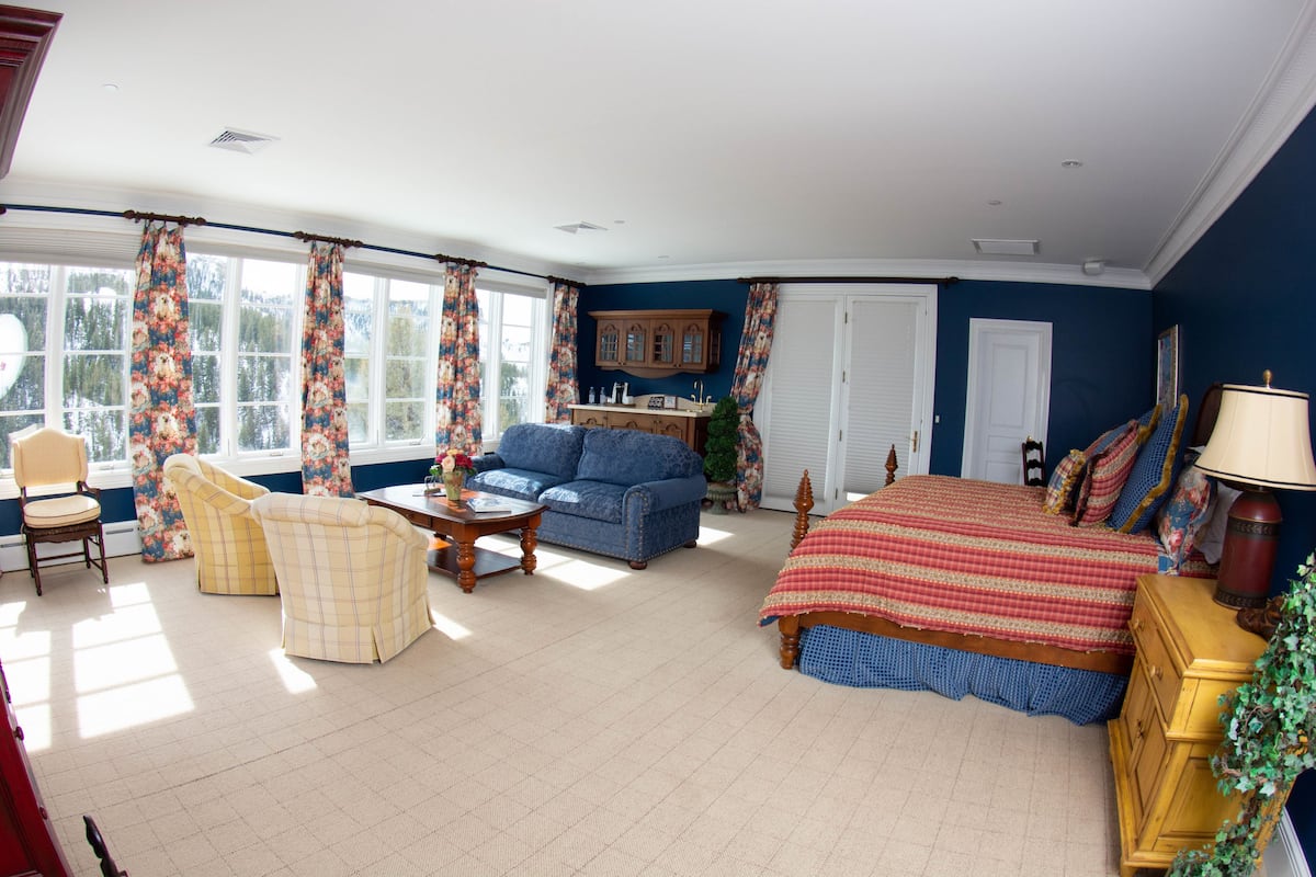 A spacious living room with a large bed, a blue sofa, and a yellow armchair. There is a wooden cabinet and a small table with a vase.