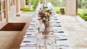 Una larga mesa de comedor dispuesta para una comida, con manteles blancos, cubiertos y centros de mesa con flores.