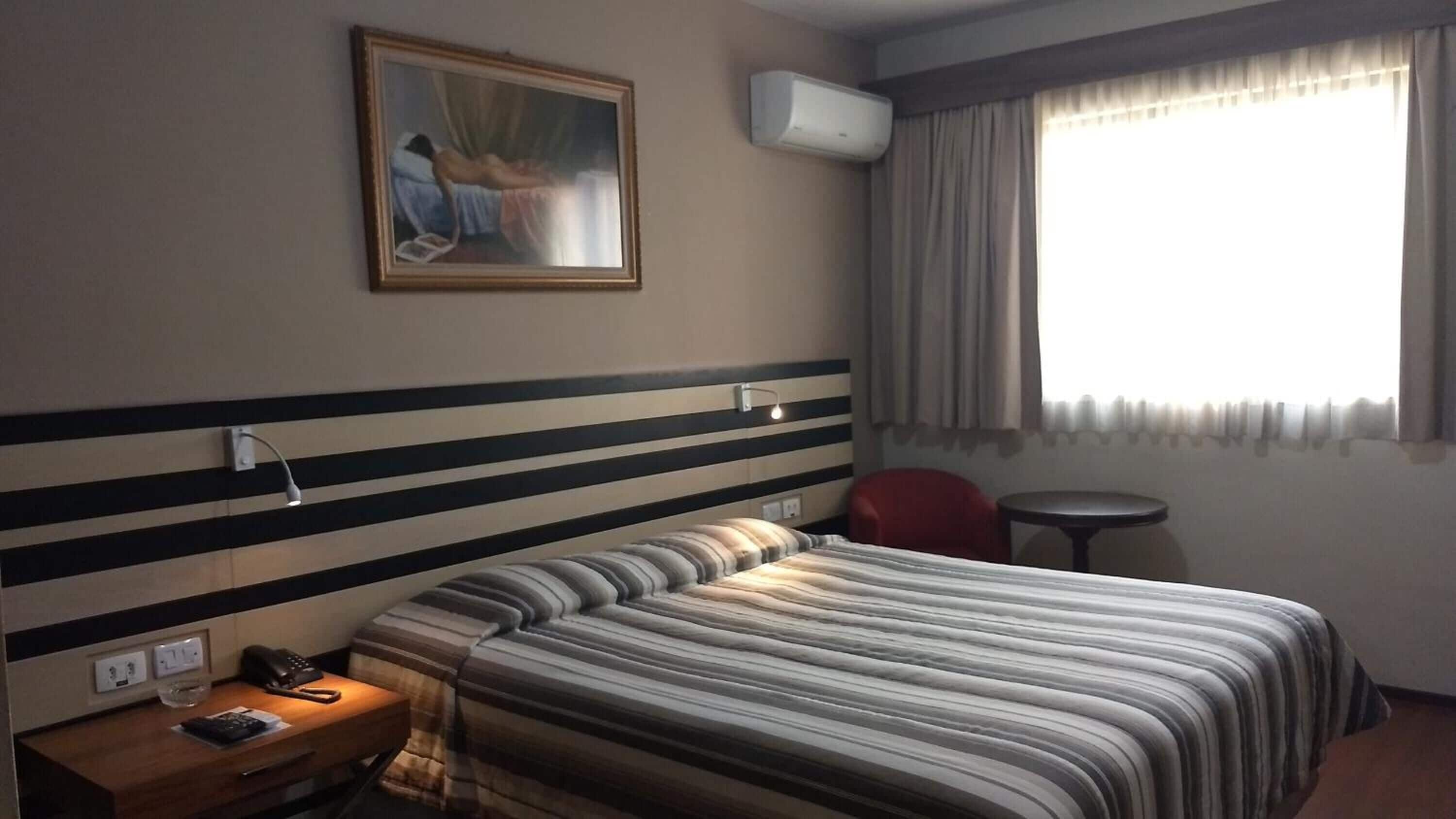 A hotel room with a bed, a nightstand, a chair, a small table, a painting, and an air conditioning unit.
