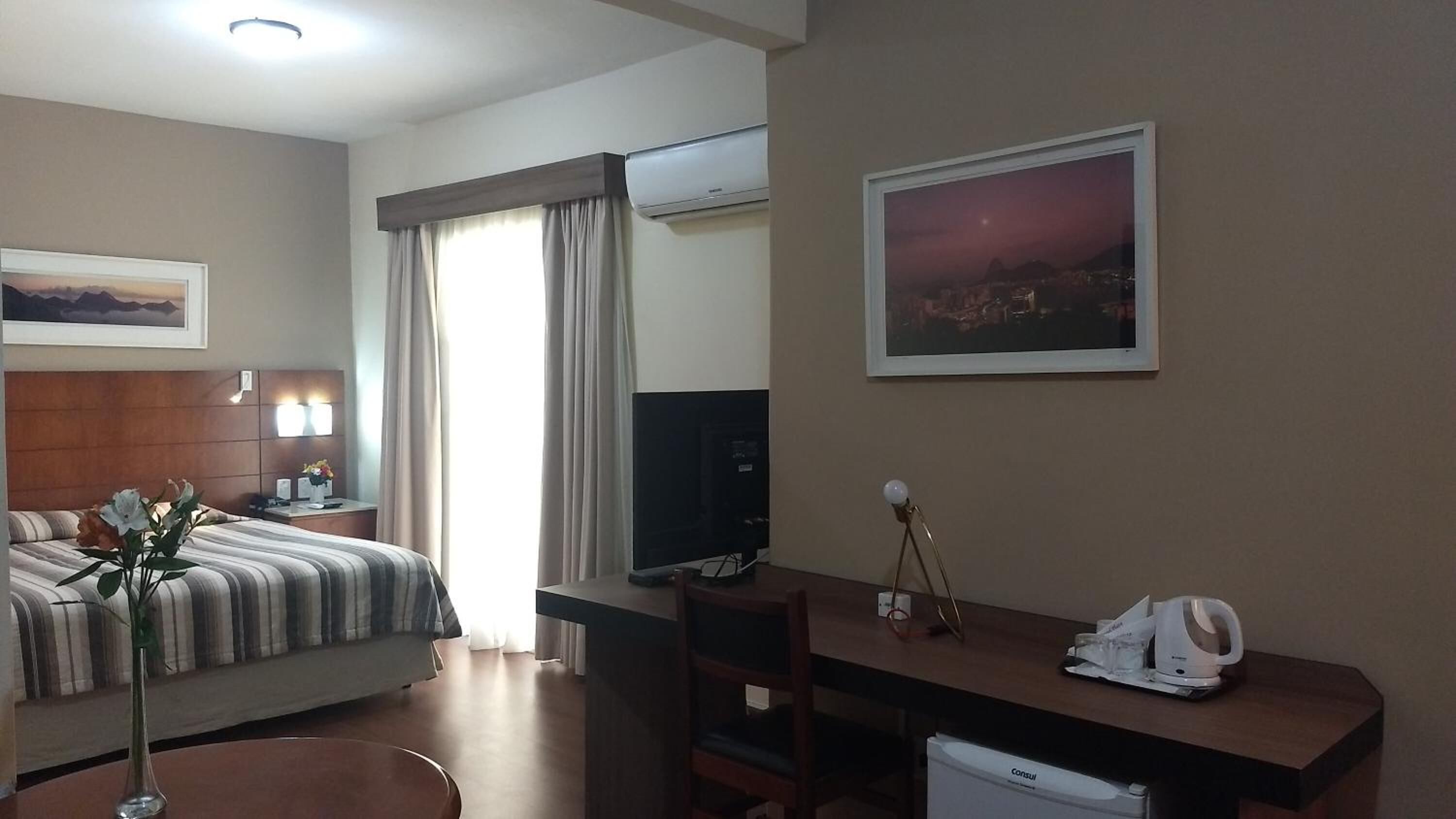 A hotel room with a bed, a desk, a chair, a television, a nightstand, a lamp, a vase with flowers, a picture on the wall, and a window with curtains.