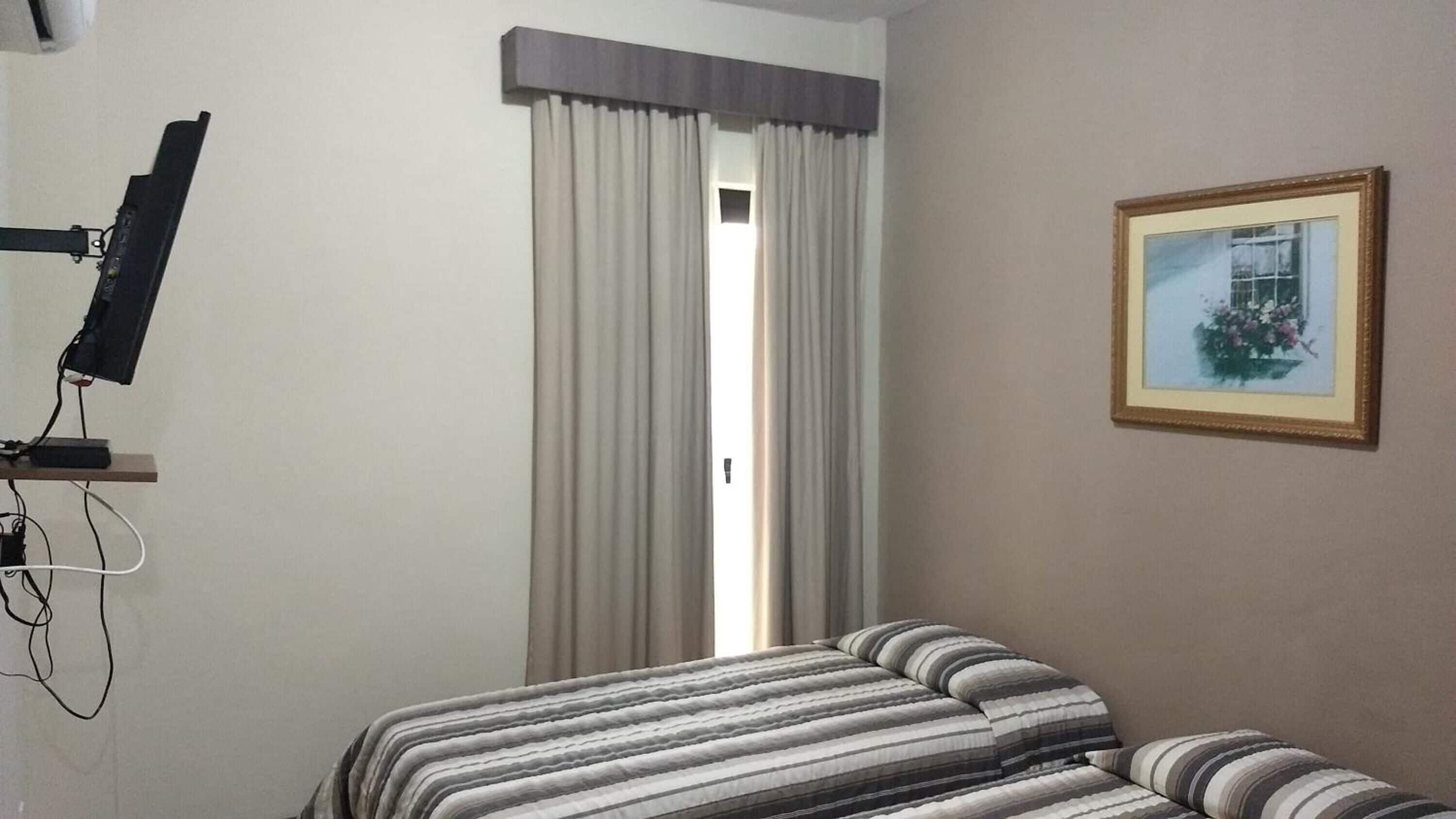 A hotel room with a bed, a television mounted on the wall, a window with curtains, and a framed picture on the wall.