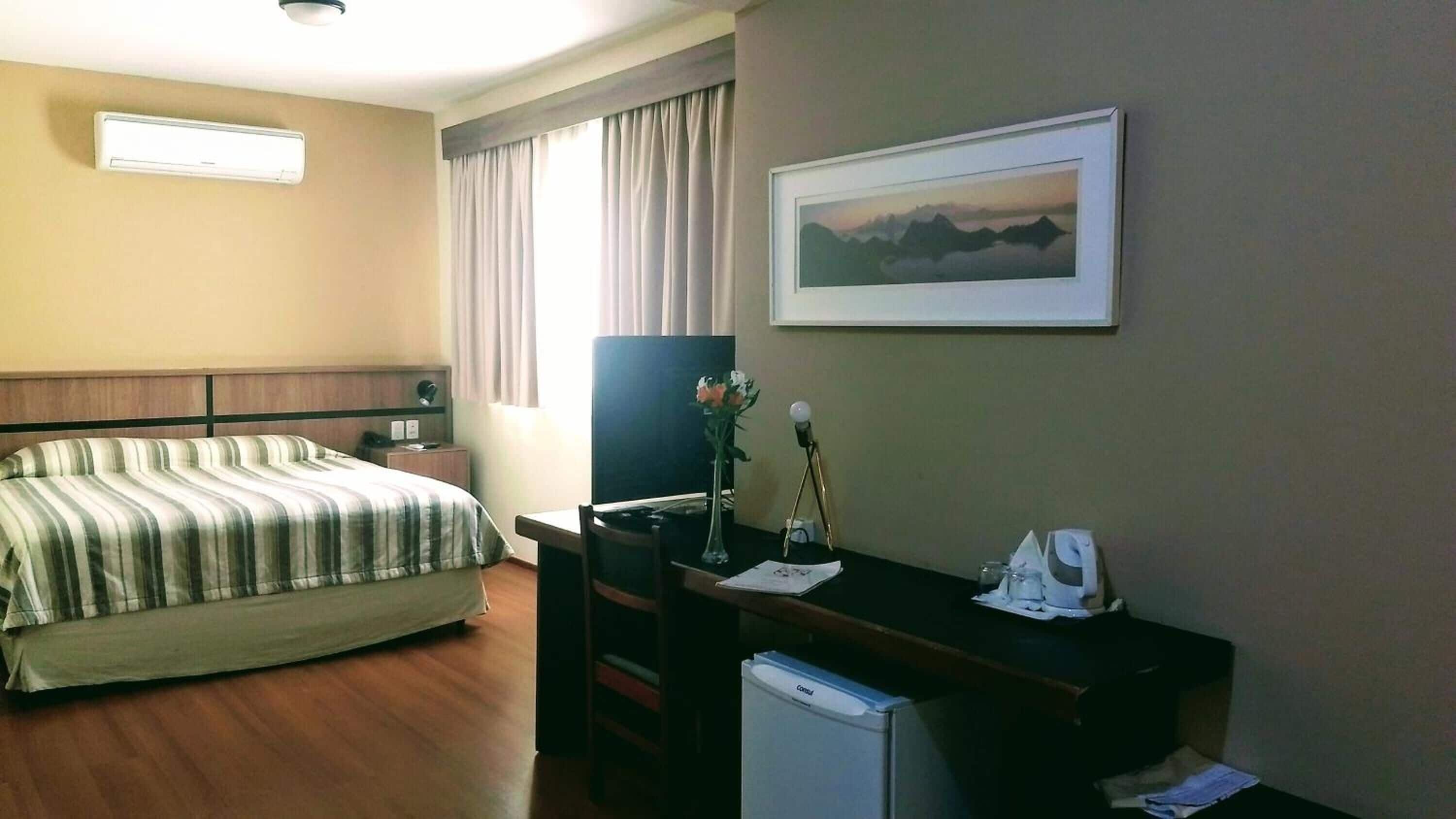 Une chambre d’hôtel avec un lit, un bureau équipé d’un ordinateur, un petit réfrigérateur, une lampe de bureau, un vase avec des fleurs, un tableau encadré au mur et une fenêtre avec des rideaux.