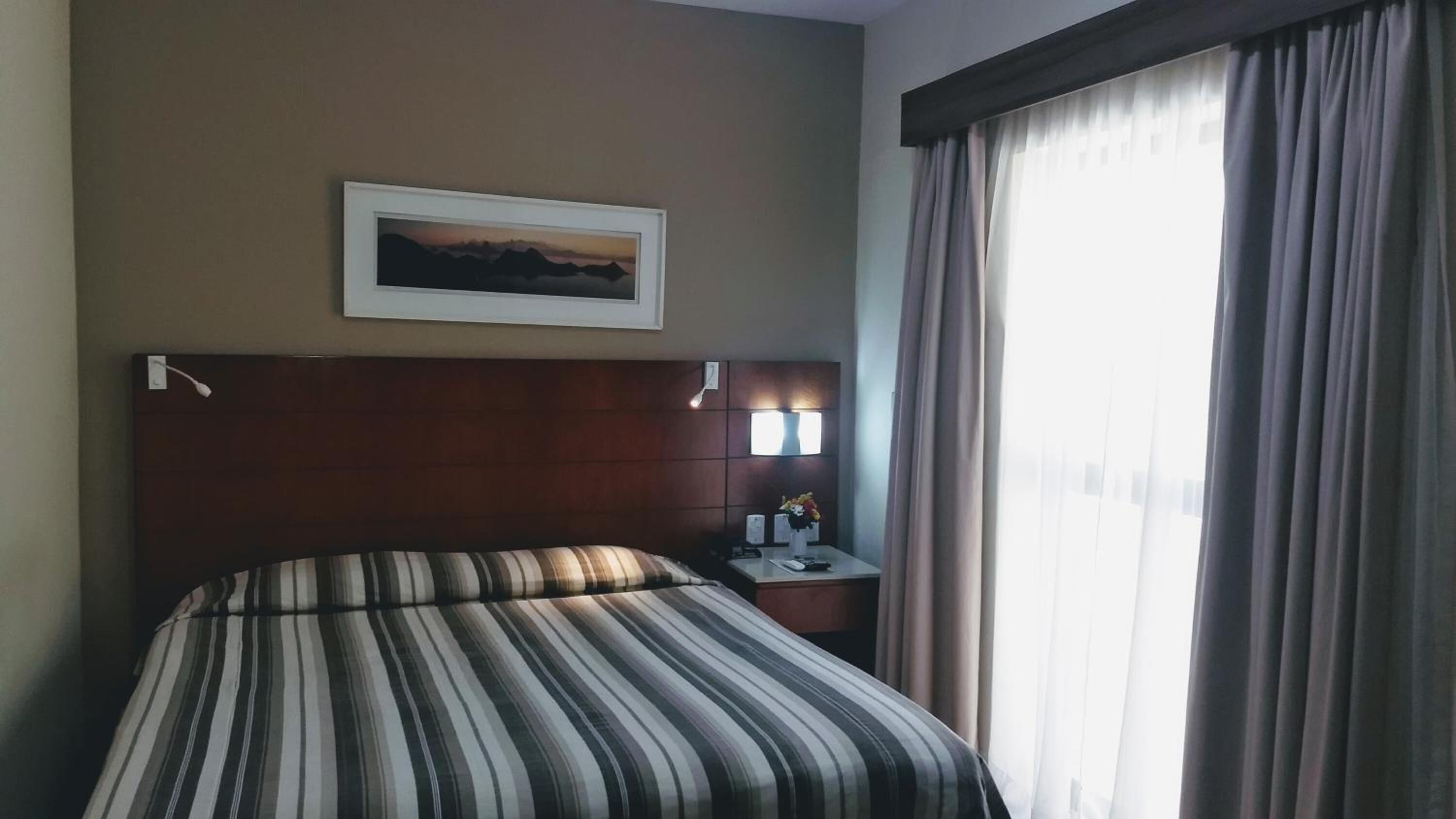 A hotel room with a large bed, a nightstand with a lamp, a framed picture on the wall, and a window with curtains.