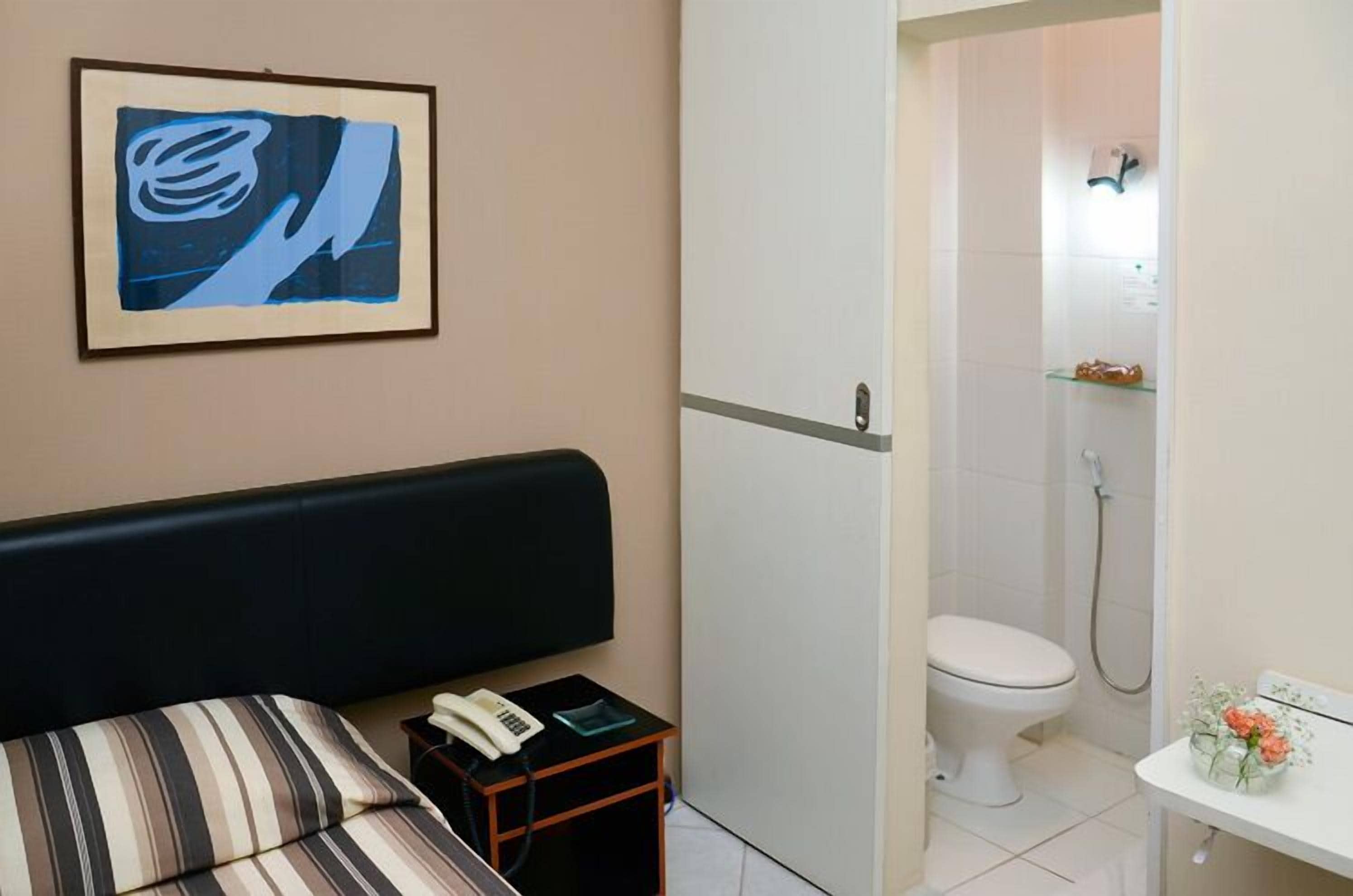 A hotel room with a bed, a bedside table with a phone, a framed artwork, a toilet, and a shower area.