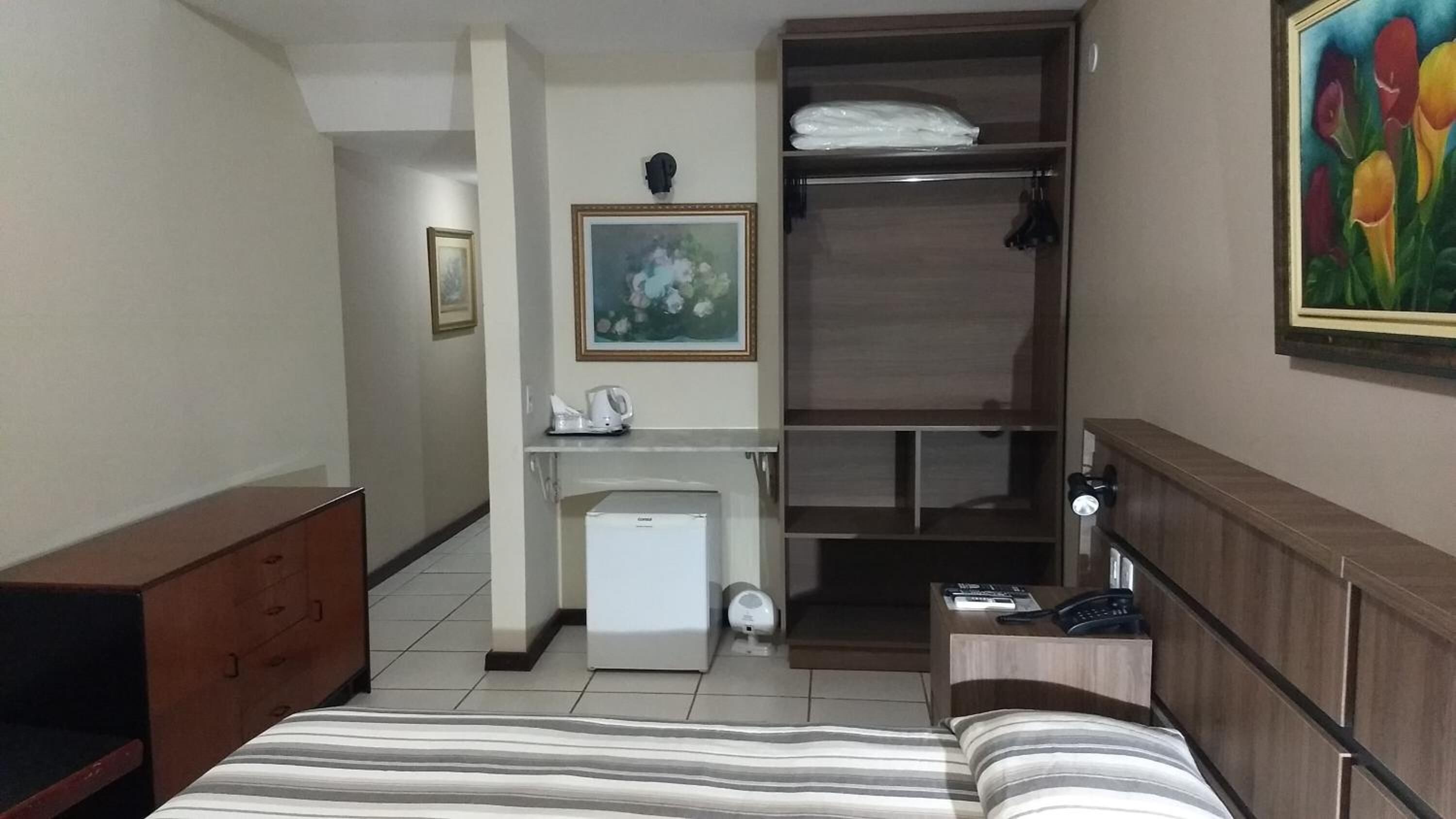 Une chambre d’hôtel comprenant un lit, un bureau avec une chaise, un petit réfrigérateur, une armoire et un tableau au mur.