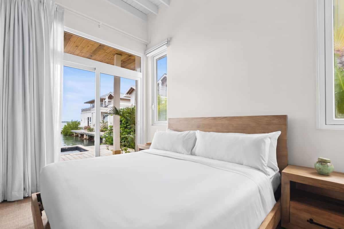 A bedroom with a large bed, wooden headboard, and a view of a pool and buildings through a glass door.
