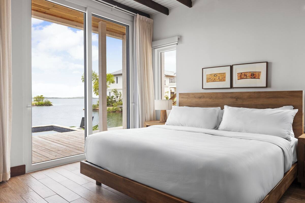 A bedroom with a large bed, a wooden headboard, a view of the water, and two framed pictures on the wall.