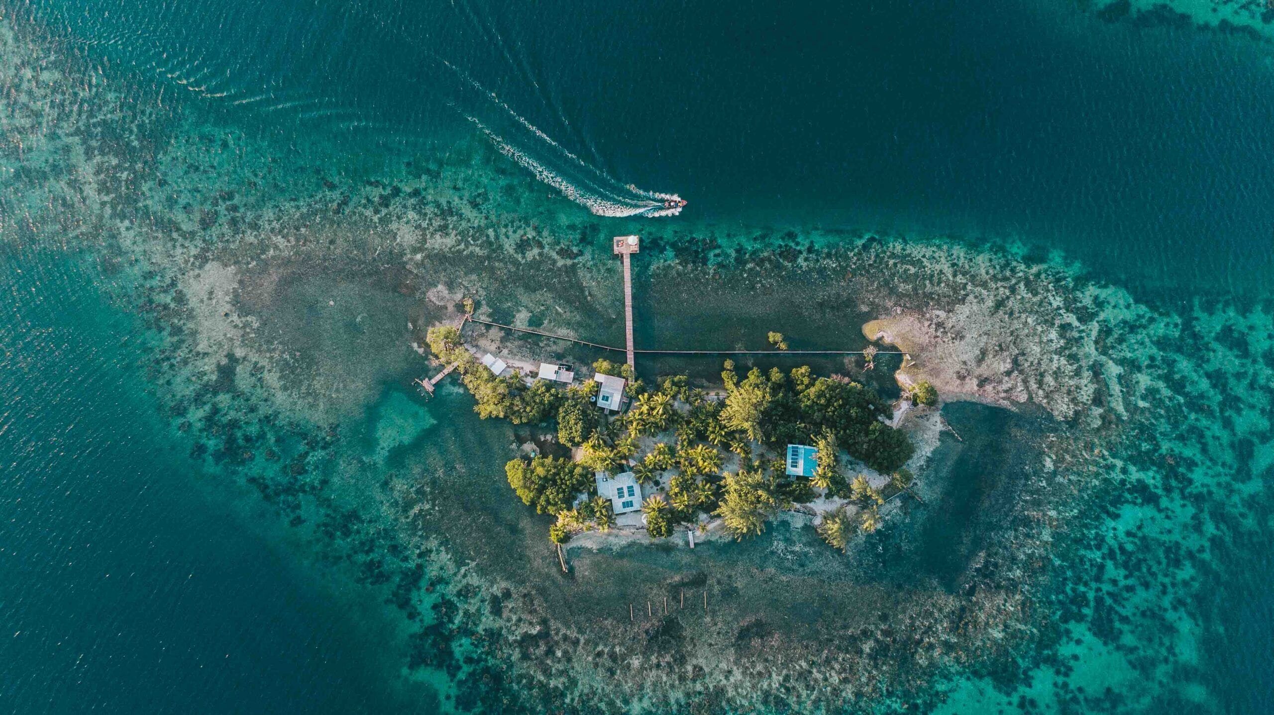Une vue aérienne d’une petite île avec un quai, quelques bâtiments et un bateau qui laisse une vague derrière lui.