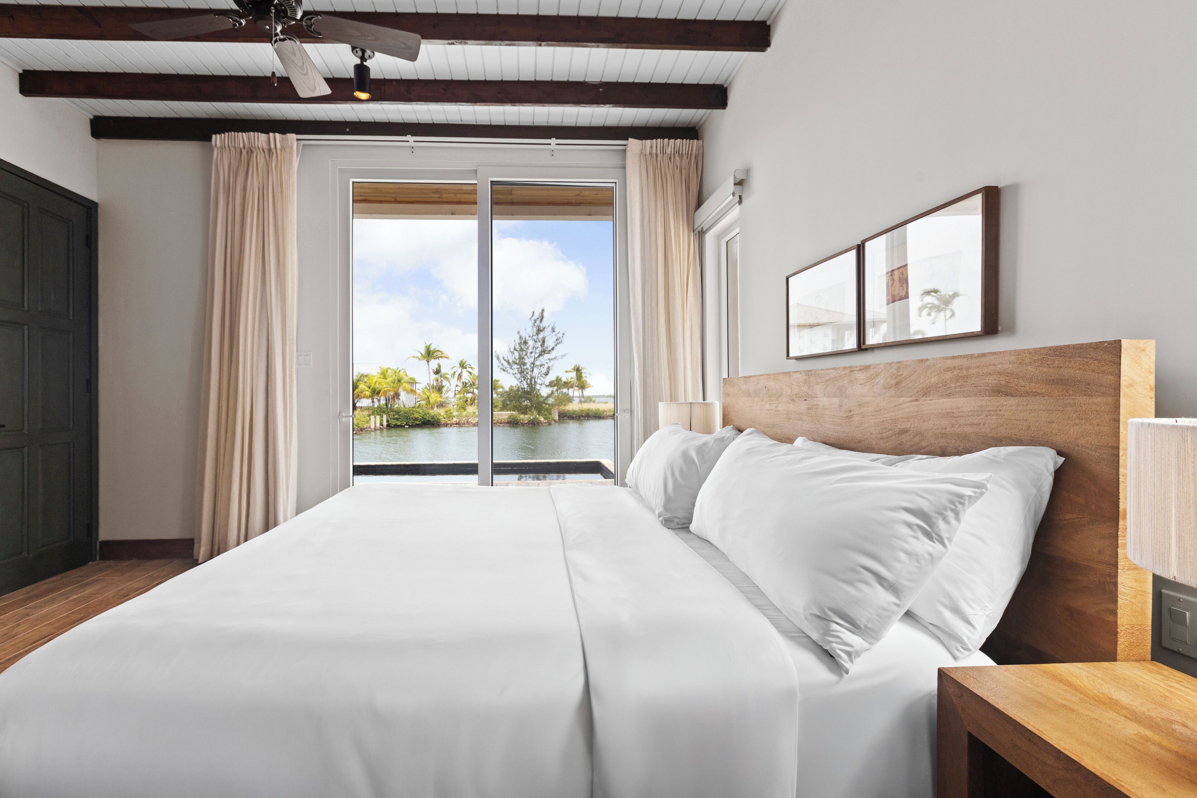 A large bed with white linens, a wooden headboard, and a view of a body of water through a sliding glass door.
