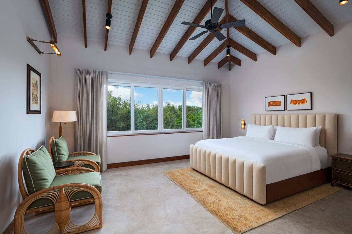 A bedroom with a large bed, a window with curtains, a ceiling fan, and a rattan chair.