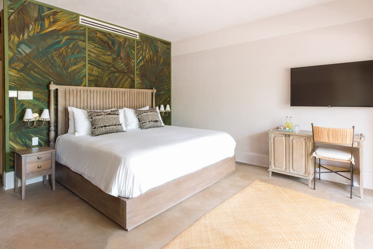 A bedroom with a large bed, a nightstand, a desk with a chair, a flat-screen TV, and a decorative wall mural.
