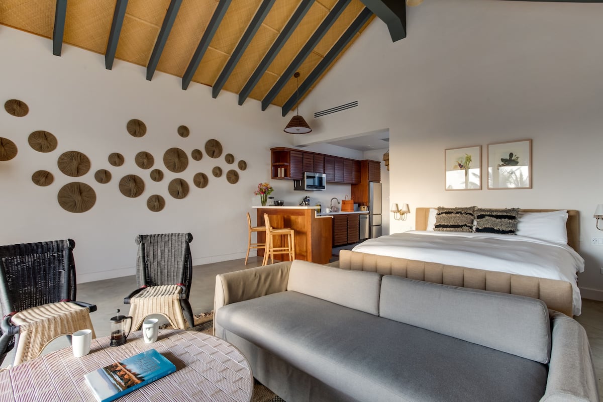 A modern hotel room with a large bed, a sofa, a dining area, and a kitchen.