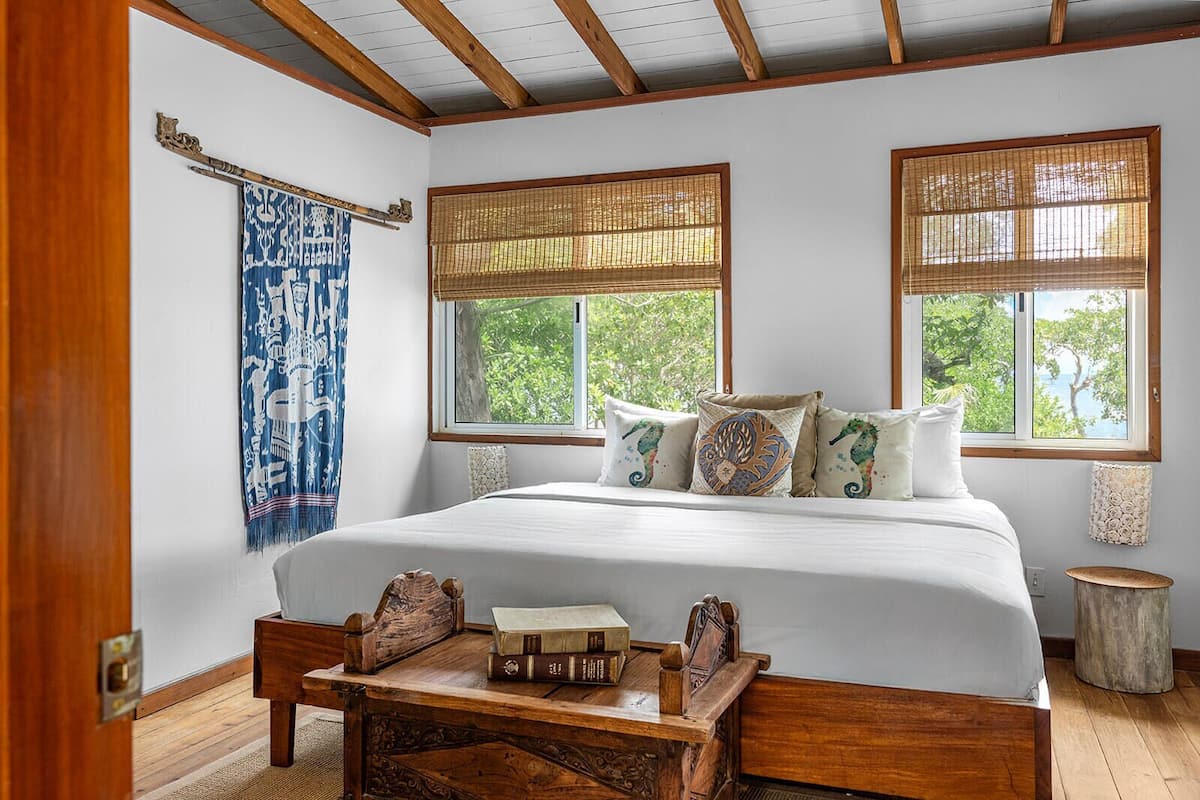A bedroom with a large bed, two windows with bamboo blinds, a wooden headboard, and a small wooden table with books.
