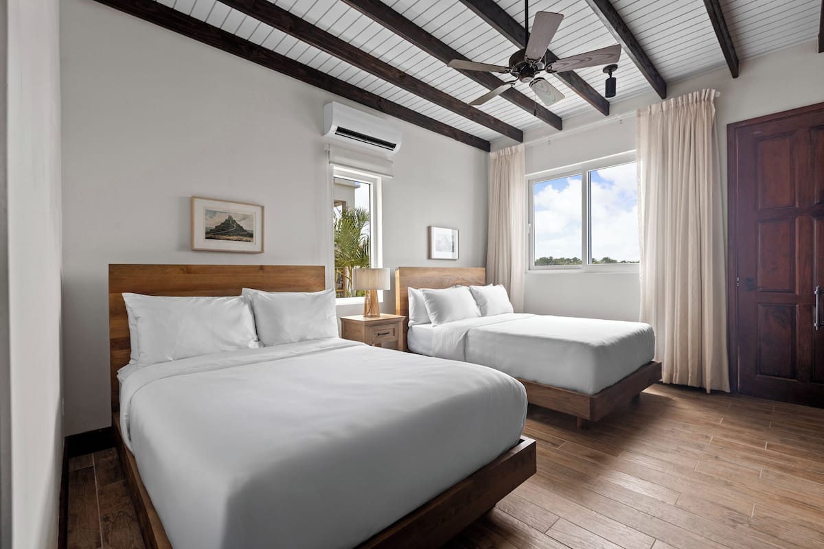 A hotel room with two beds, wooden headboards, a ceiling fan, and a large window with curtains.
