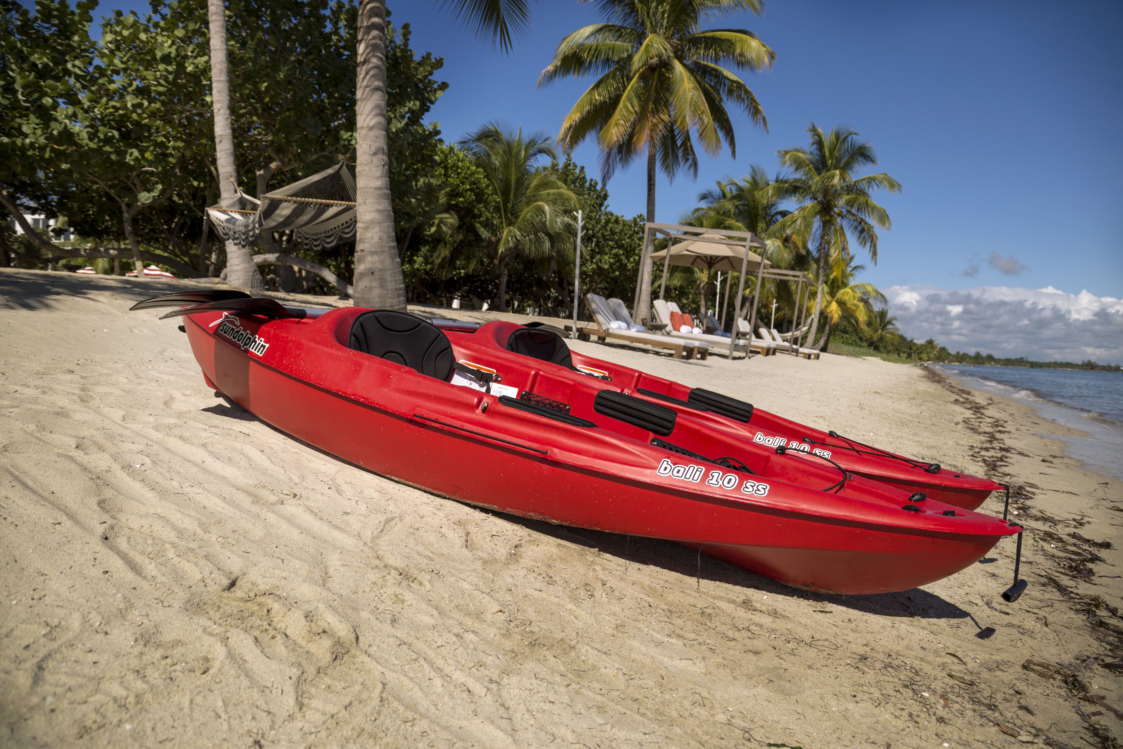 A red kayak with the inscription "bali 10 ss" on a sandy beach with palm trees and a clear sky.
