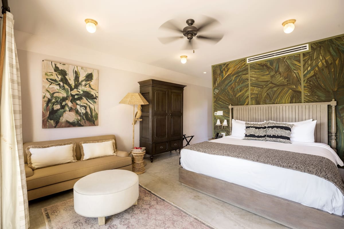 A bedroom with a bed, a sofa, a ceiling fan, and a large painting on the wall.