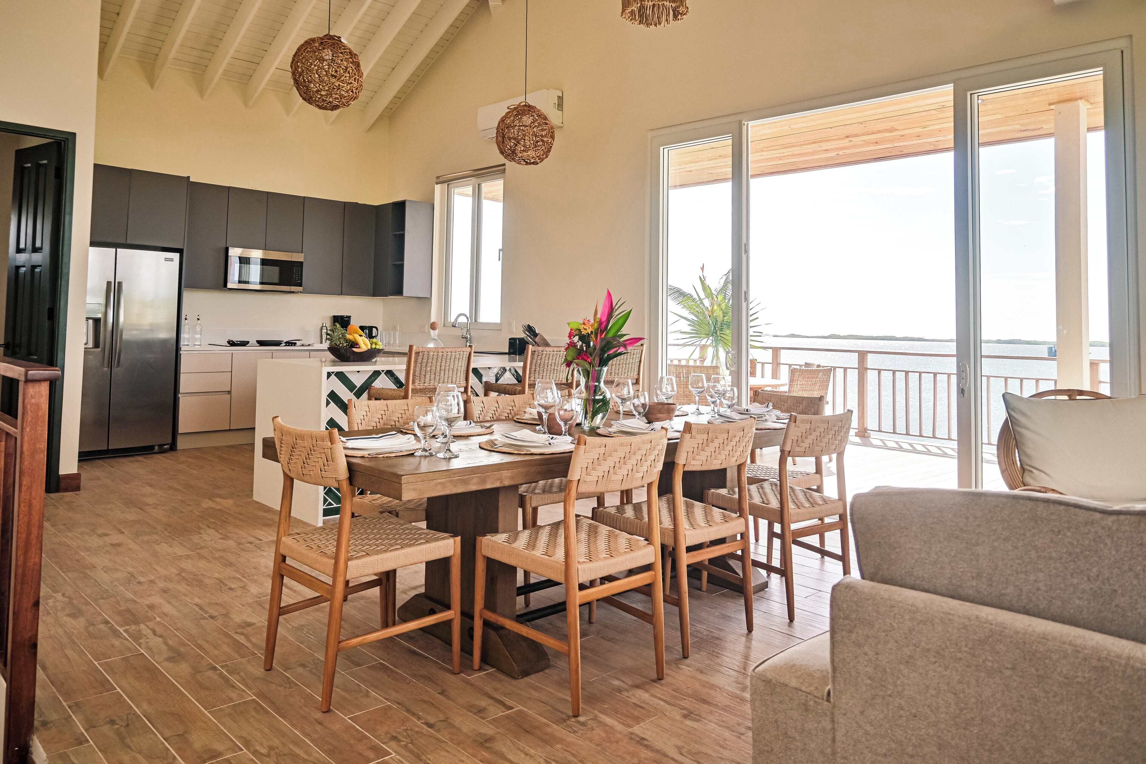 Une vaste salle à manger avec une table en bois dressée pour six personnes, entourée de chaises en osier. On aperçoit une cuisine équipée d’appareils modernes au fond, et un balcon avec vue sur la mer est également présent.
