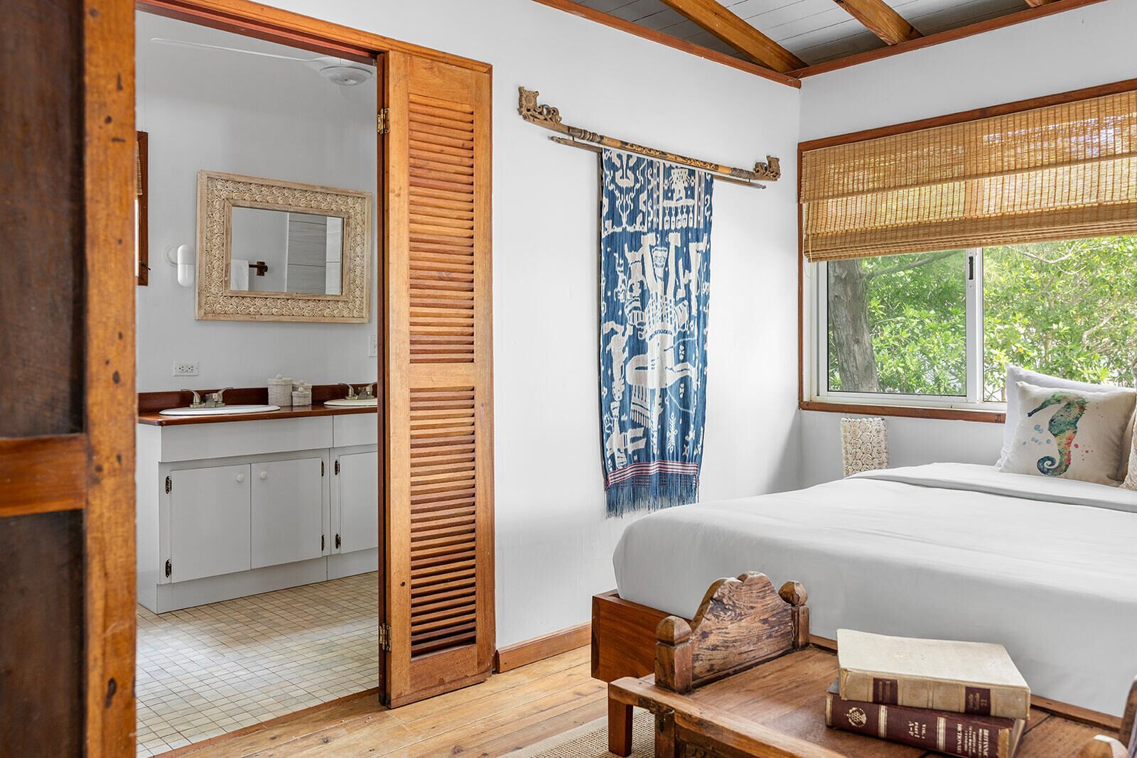Un dormitorio con cama de madera, ventana con vistas, mesita de noche con libros y baño con espejo y lavamanos.