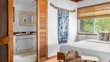 Un dormitorio con cama de madera, ventana con vistas, mesita de noche con libros y baño con espejo y lavamanos.