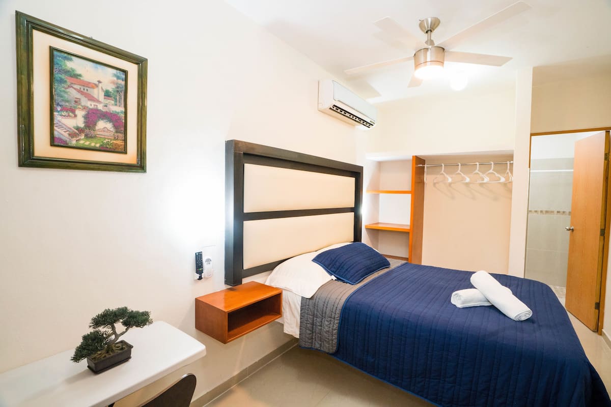Ein Schlafzimmer mit einem Bett, einem Nachttisch, einer kleinen Pflanze, einem Deckenventilator und einer Wandklimaanlage.