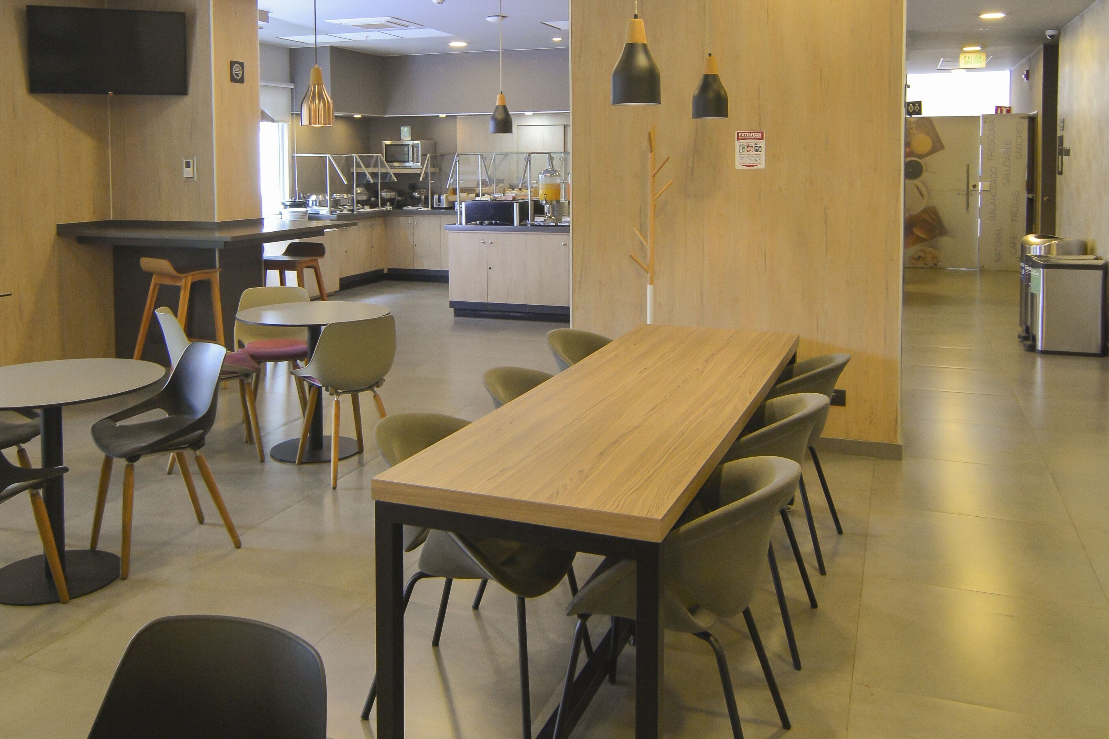 A modern dining area with a long wooden table and several chairs, a kitchen in the background, and pendant lighting.