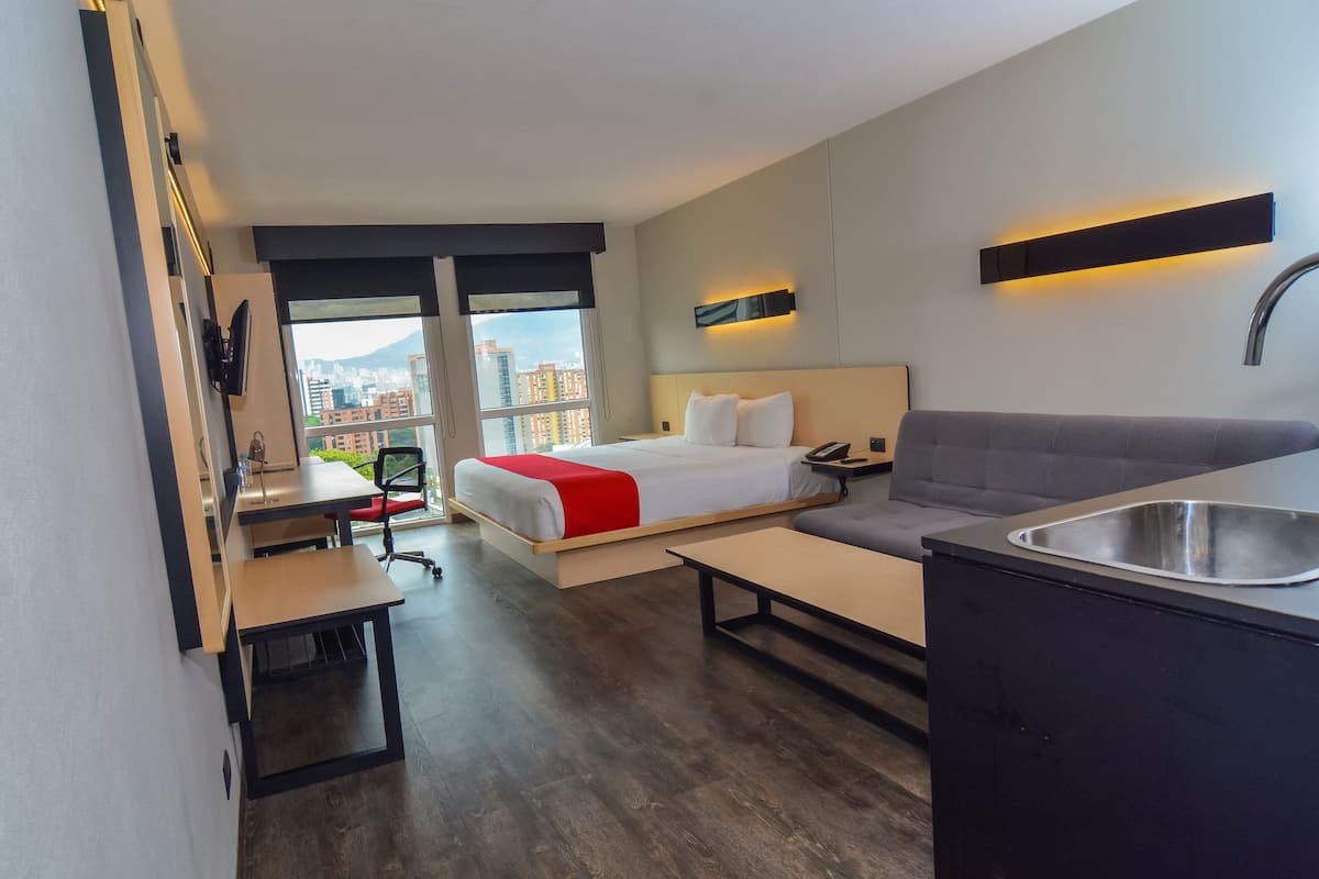 A modern hotel room with a bed, a sofa, a desk, and a kitchenette.