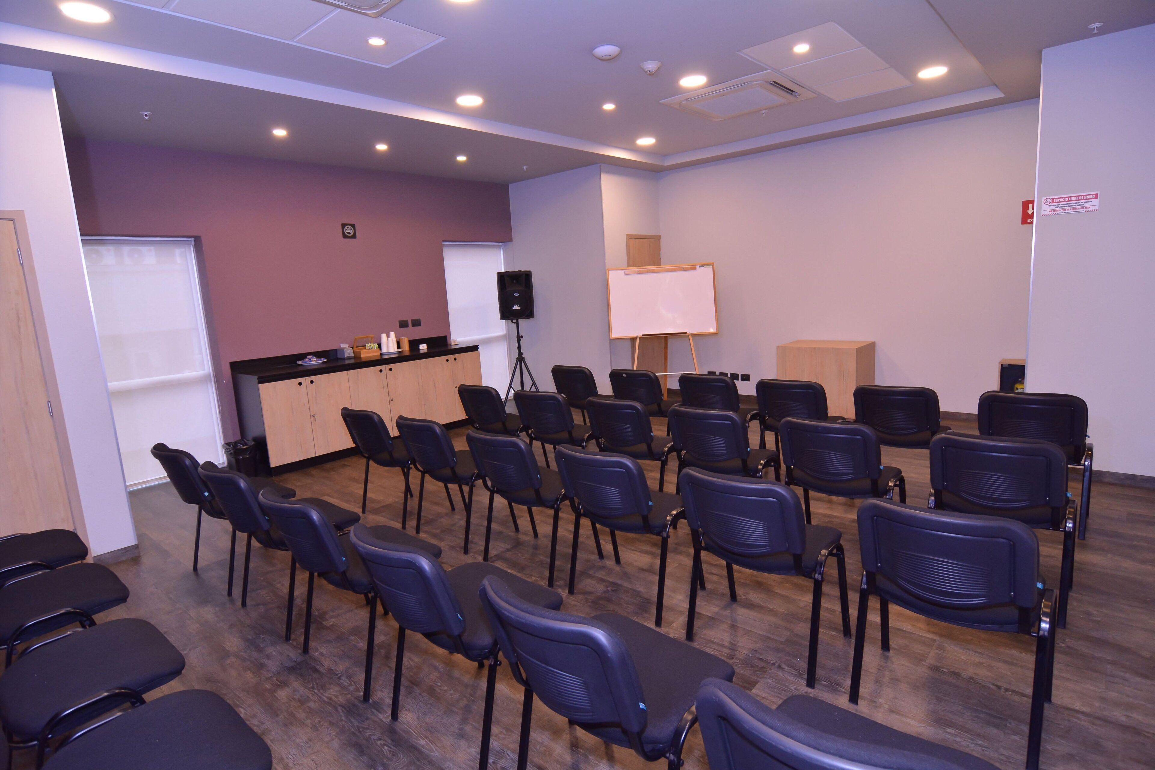 Un salón de conferencias con filas de sillas, un atril, una pizarra blanca y un altavoz sobre un trípode.