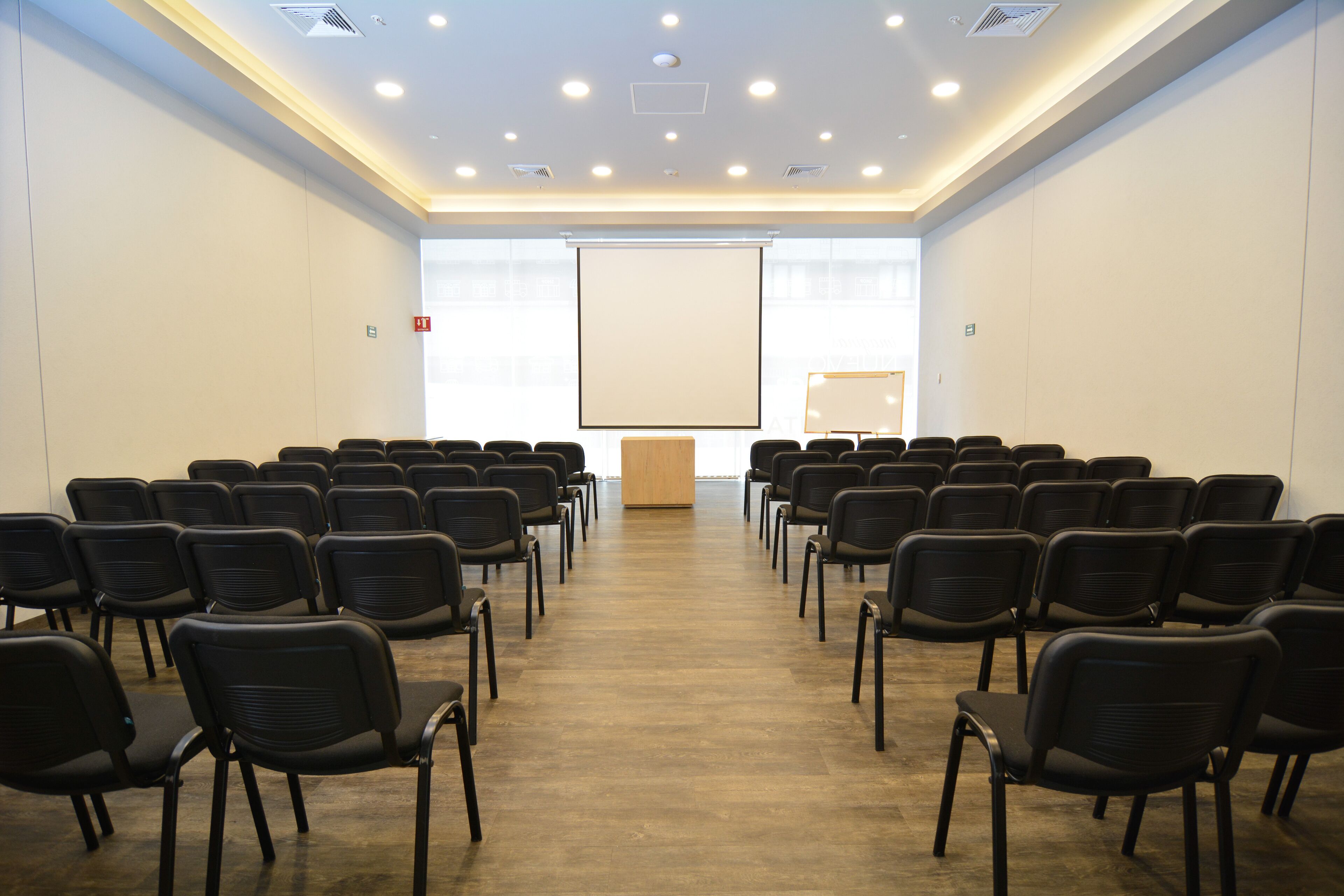 Un salón de conferencias con filas de sillas negras, un atril y una pantalla de proyección grande.